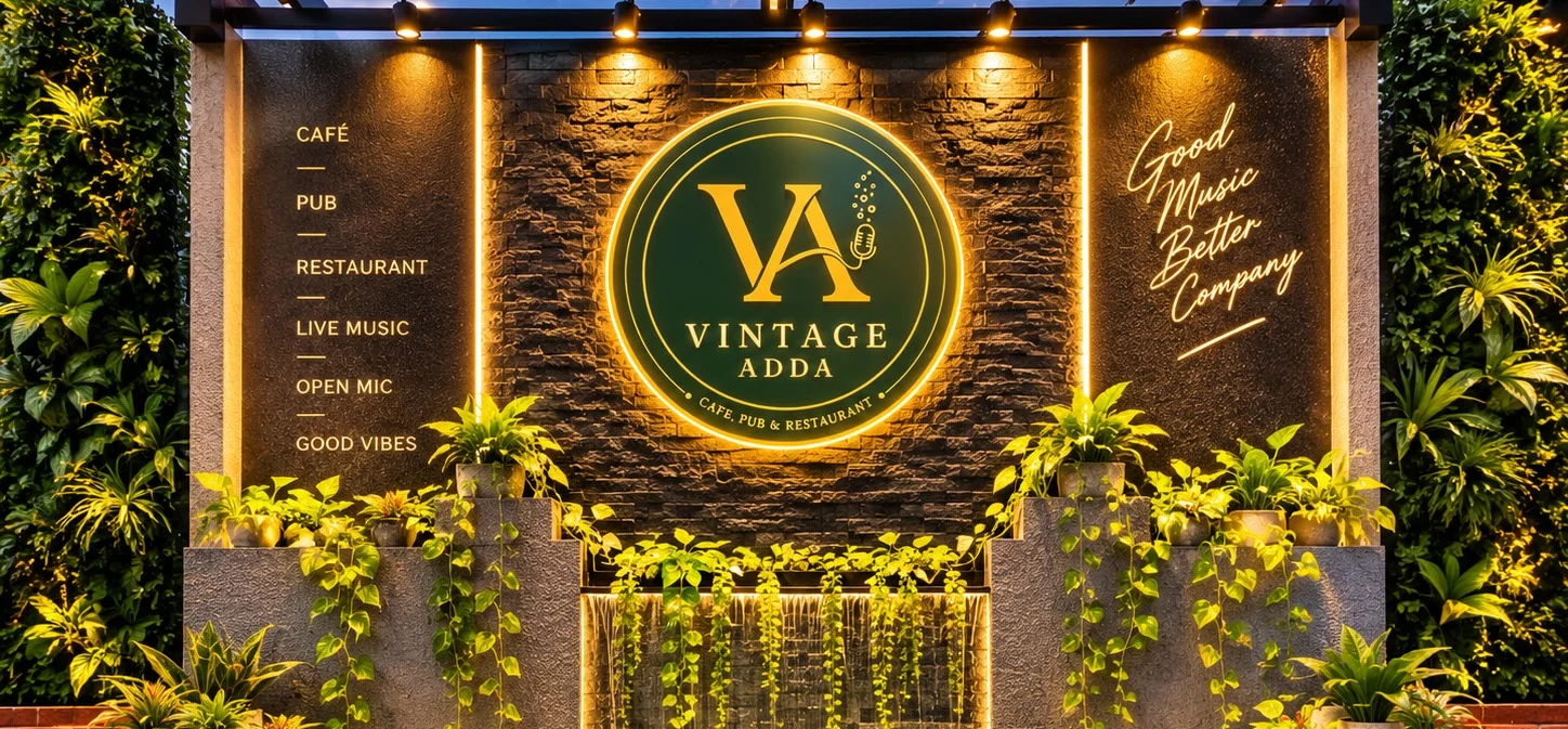The lit feature wall at the entrance to Vintage Adda: the backlit VA badge on stone, the venue’s list beside it, and a low waterfall running through banked planting below.
